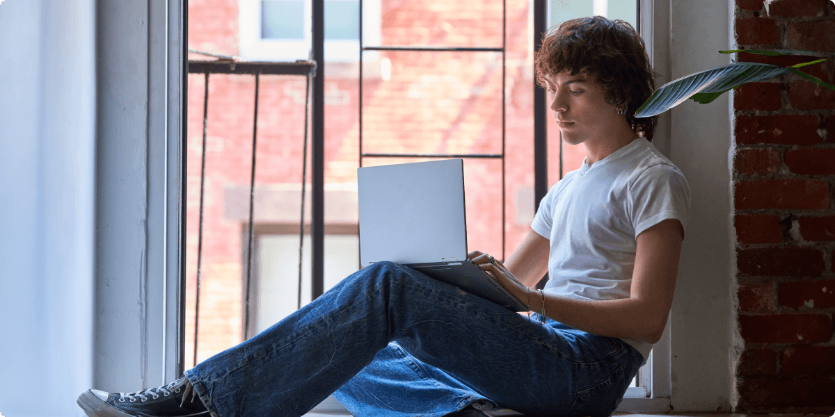 Ein junger Mann sitzt lässig auf einem Fensterbrett mit seinem Laptop.