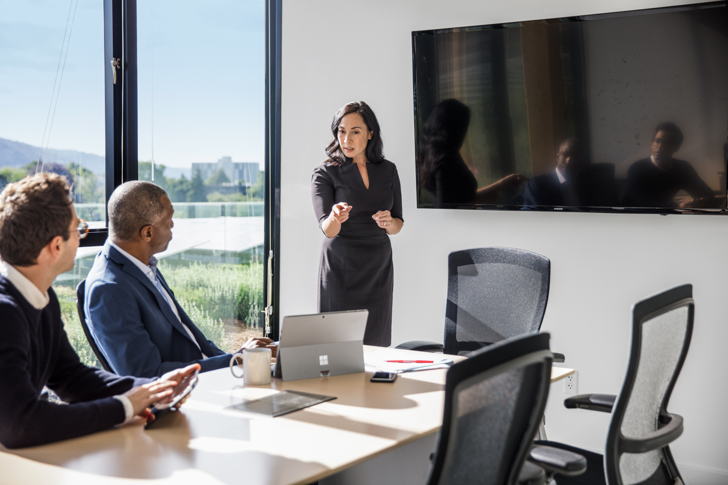 The office workers meet in a small conference room. One gives a presentation.