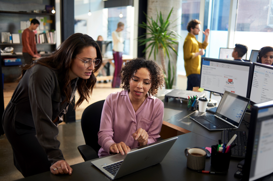 Two women looking at a laptop computer in a busy office.