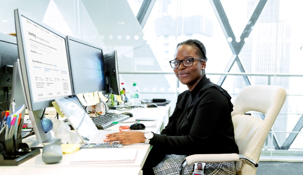 Woman at a desk who is looking towards a camera set in front of computer monitors.