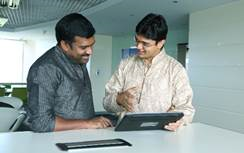 Two men standing at a counter smiling and looking at a tablet computer one of the men holds.