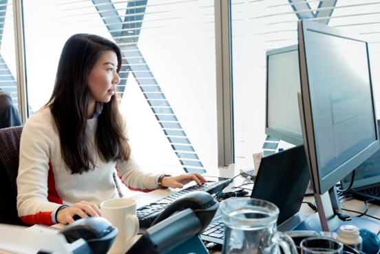 Woman near a window smiling and looking at two computer monitors.