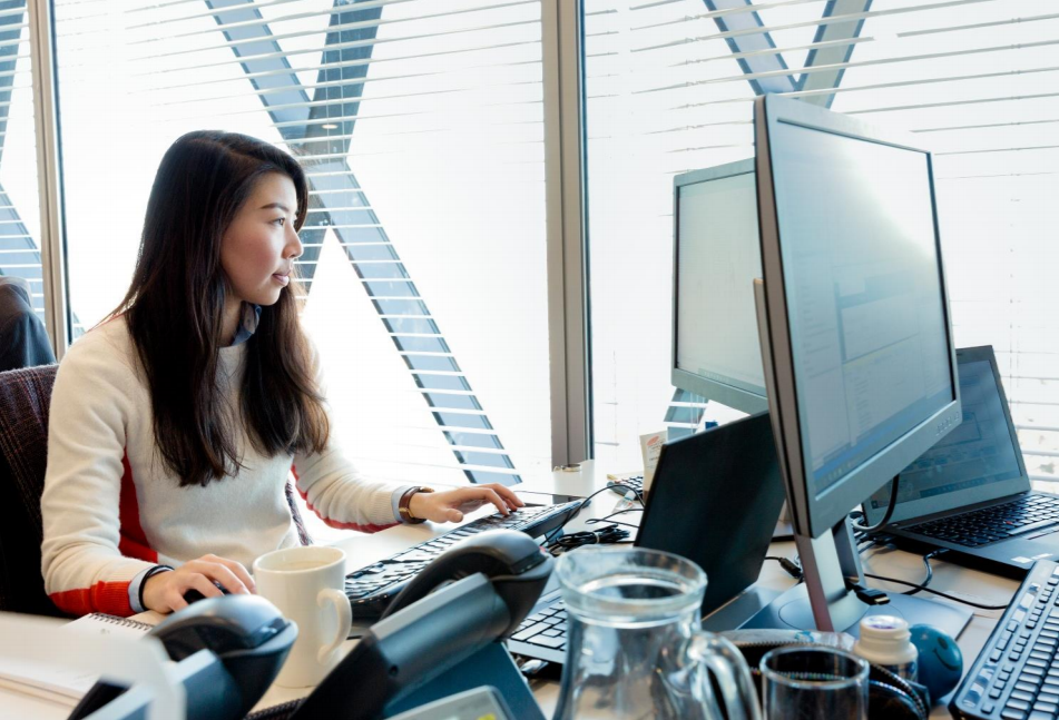 Woman sitting at an office desk in front of a laptop and monitor.