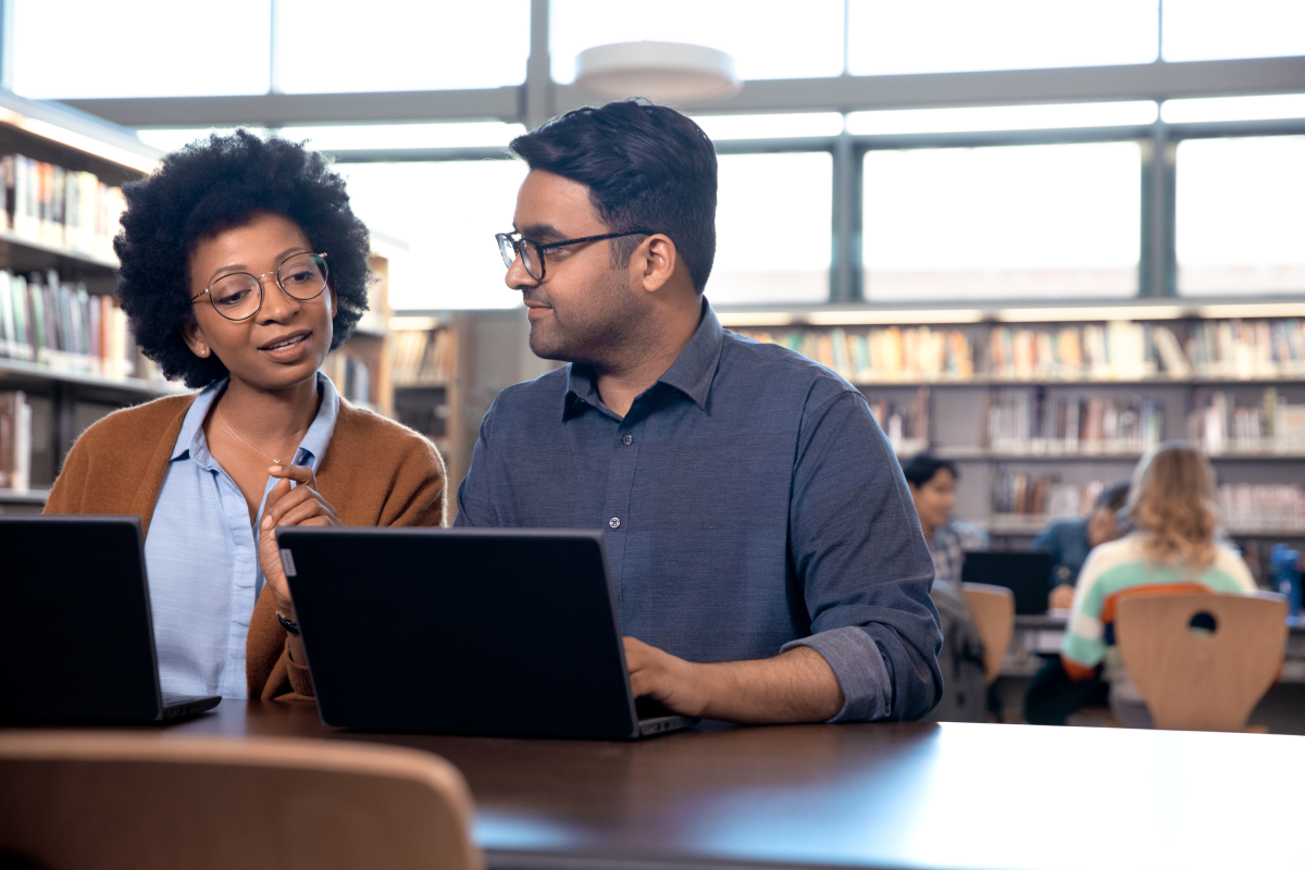 Photograph of two adults working on laptops and speaking to each other in a library.