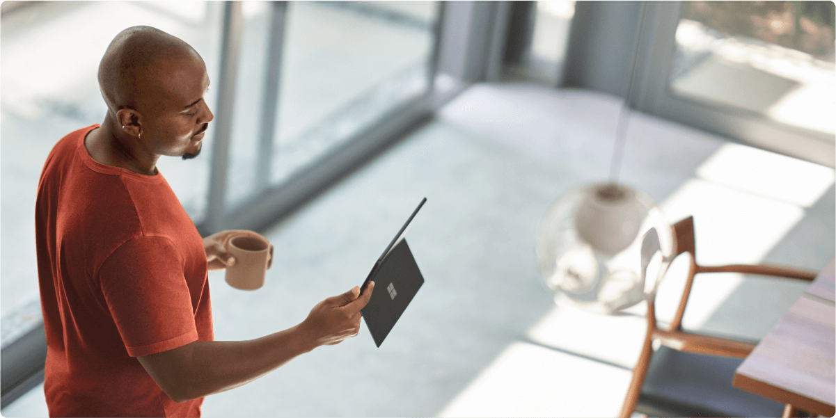 Un hombre mirando una tableta mientras disfruta de su café matinal. La luz del sol entra por las ventanas de su casa.