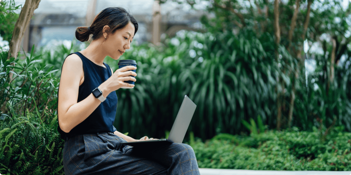 Donna al computer all'esterno con una tazza di caffè. È circondata da una vegetazione rigogliosa.