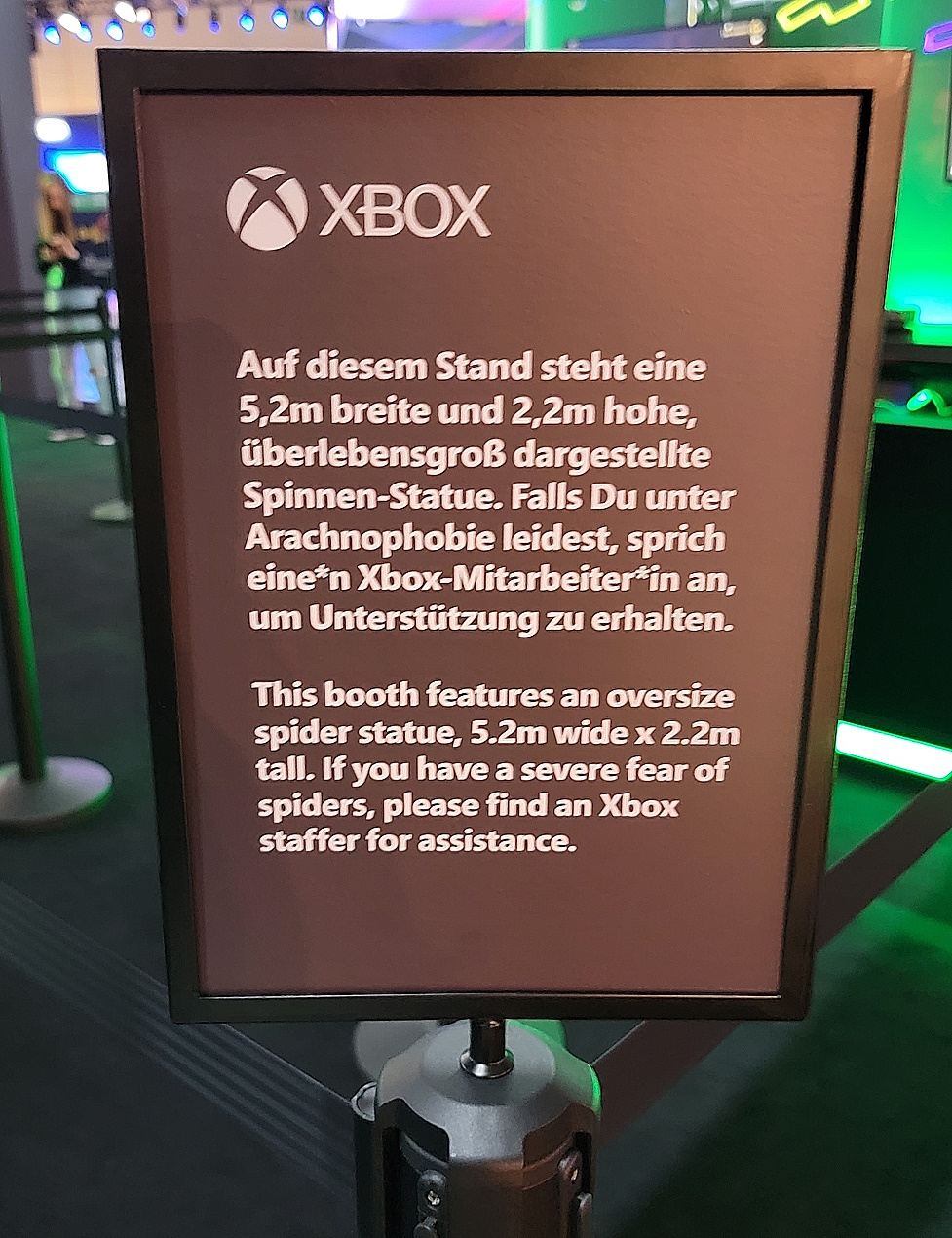 英語とドイツ語の両方で読む白いテキストを持つ黒い看板「このブースには、幅5.2m×高さ2.2mの大型スパイダー像があります。クモに対する深刻な恐怖がある場合は、Xbox スタッフにお問い合わせください。