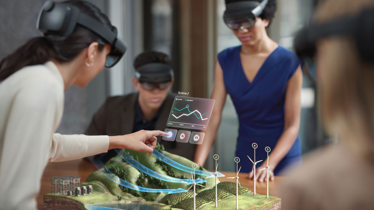 Foto van meerdere gebruikers die HoloLens-apparaten dragen en interactie hebben met een meeslepende ervaring op een tafel met terrein met windturbines.