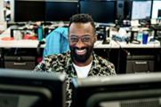 Foto de um homem sorrindo e olhando para dois monitores de computador.