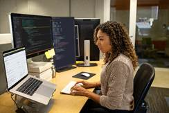 Mulher sentada e programando em sua mesa em um laptop com vários monitores de computador grandes.