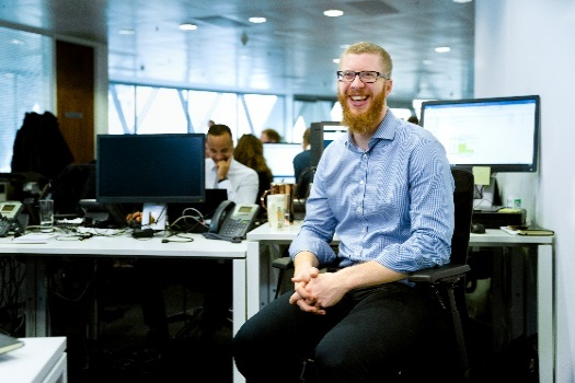 Foto de um homem sorrindo para uma câmera em um escritório movimentado.