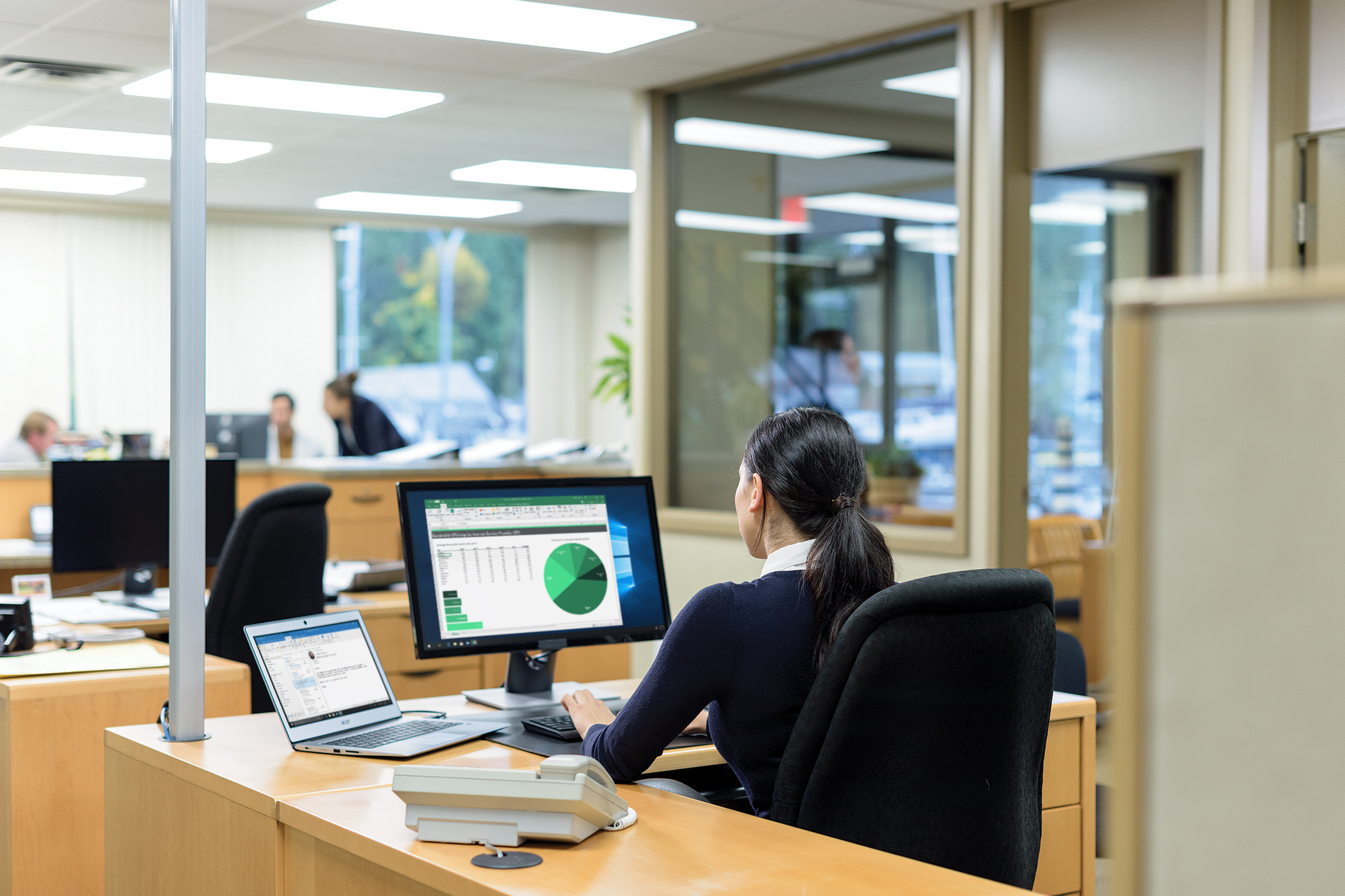 An office worker views a chart and tables on a screen while others meet in the background.